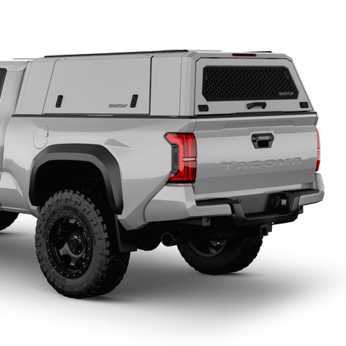 Rear three-quarter view of a white Toyota Tacoma pickup truck with a black Smart Cap canopy cover on the bed, black rear bumper and diffuser, and black off-road tires on a white studio background.
