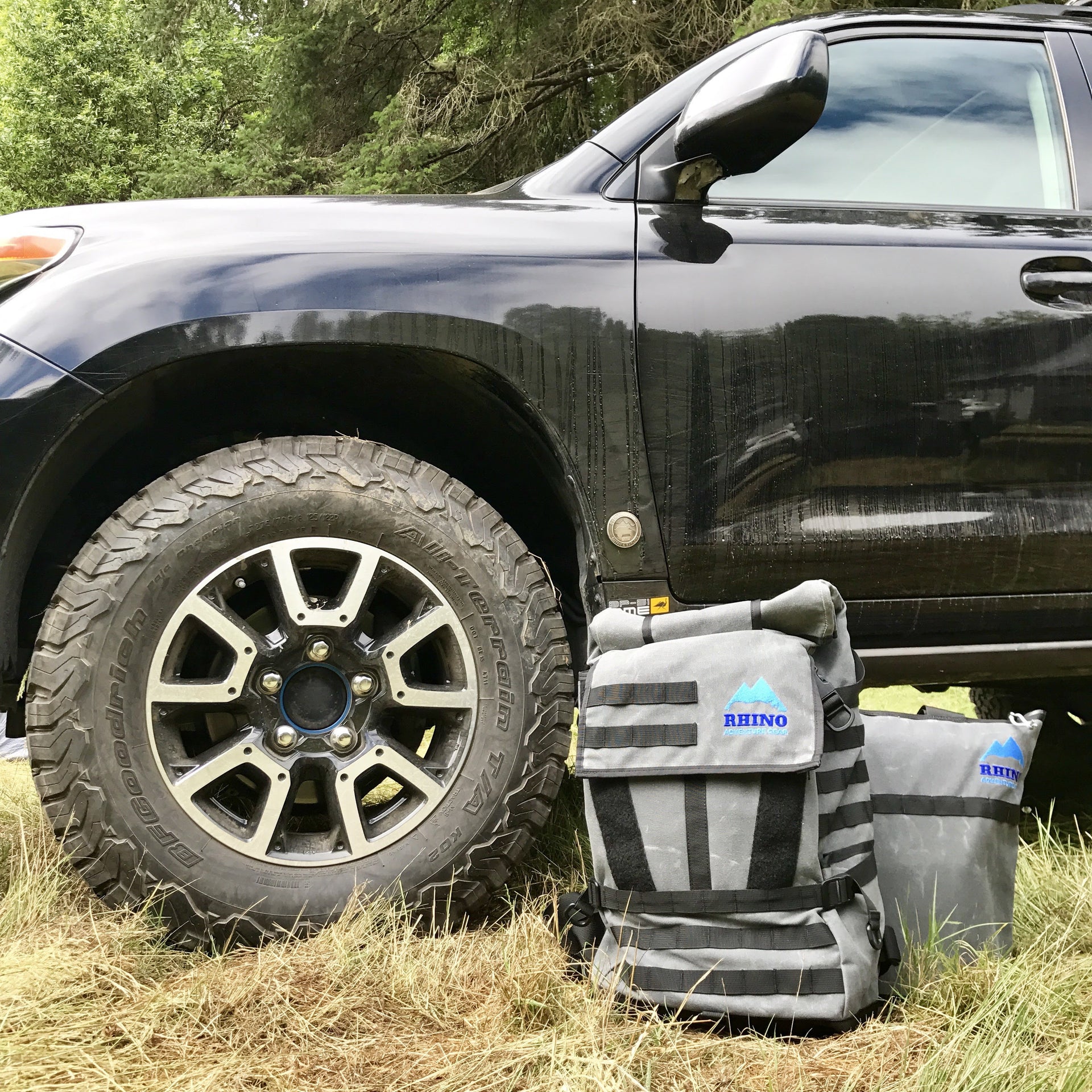 Adventure Backpack and Adventure Tote Bag with Rhino Adventure Gear embroidered logo leaning against black Toyota Landcruiser near wheel