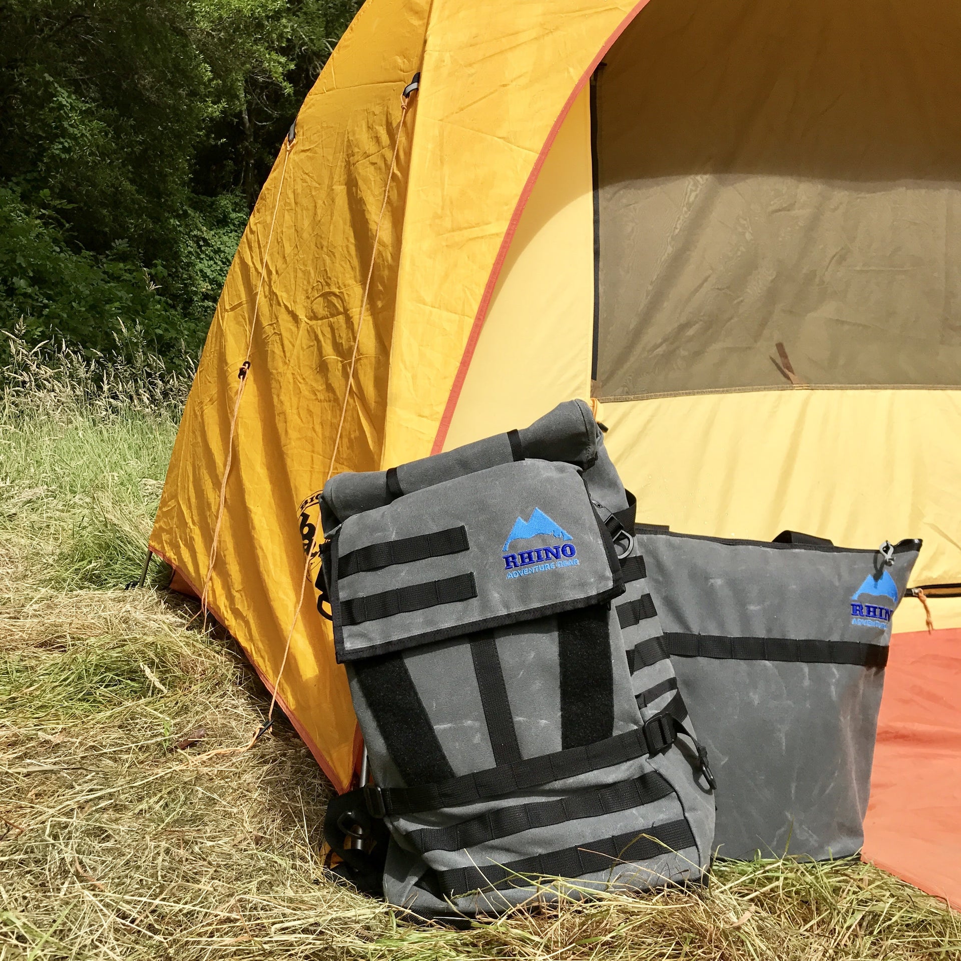 Adventure Tote Bag and Adventure Backpack, both grey with black trim and blue Rhino Adventure Gear logo shown leaning against yellow and orange tent