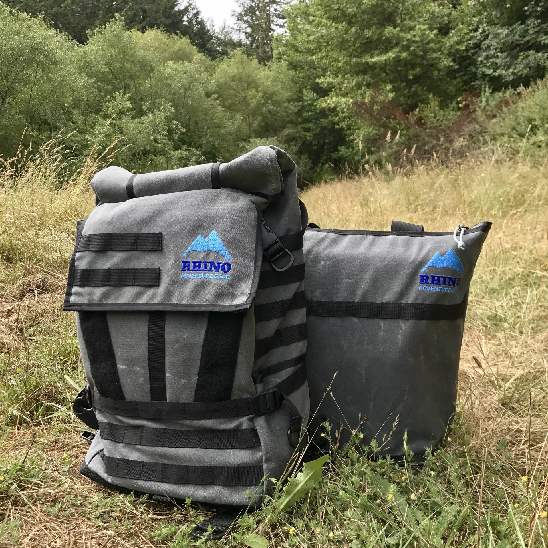 Adventure Tote Bag and Adventure Backpack, both grey with black trim and blue Rhino Adventure Gear logo shown sitting in grassy field