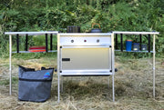 Trail Kitchens Camp Kitchen with Integrated Stove set up outdoors viewed head-on showing stove knob controls and shelving hanging below counters for additional storage